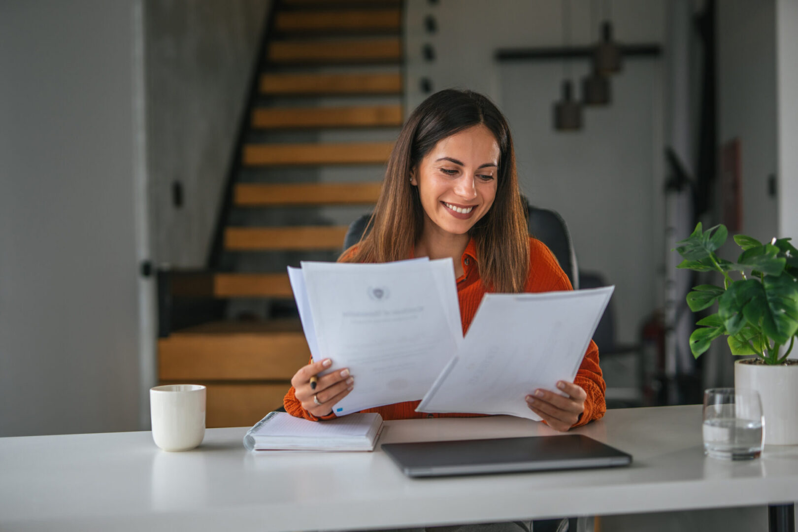 Young woman working productively and happily from a home office, sitting at a white desk and reviewing important paperwork while holding documents, embodying remote work and success concepts